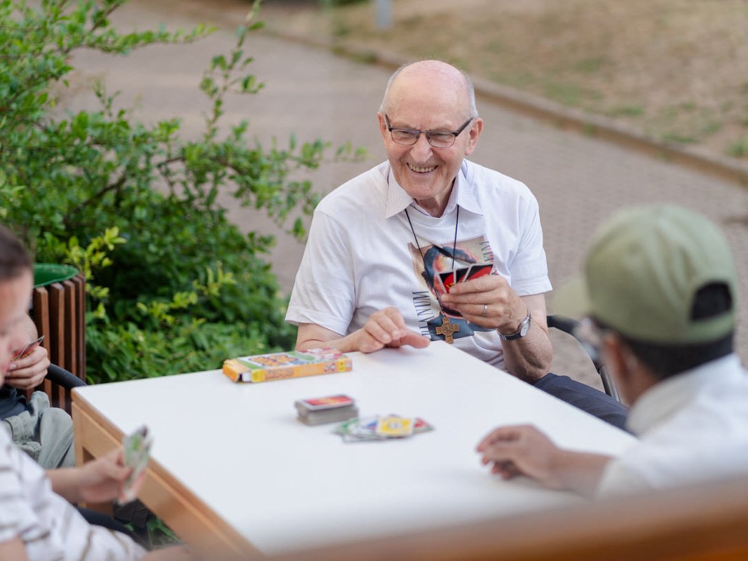 Glücklicher Senior mit Brille spielt draußen Karten mit Freunden und lächelt breit.