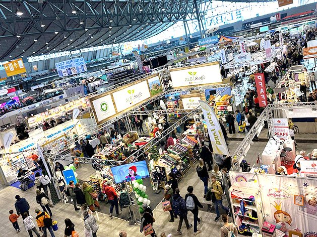 Blick von oben auf eine belebte Messehalle voller Ausstellerstände und Besucher, die Stoffe und Bastelbedarf ansehen.