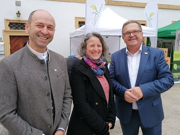 Drei lächelnde Personen, zwei Männer und eine Frau, stehen im Freien vor Zelten eines Natur- und Forstwirtschaftsevents.