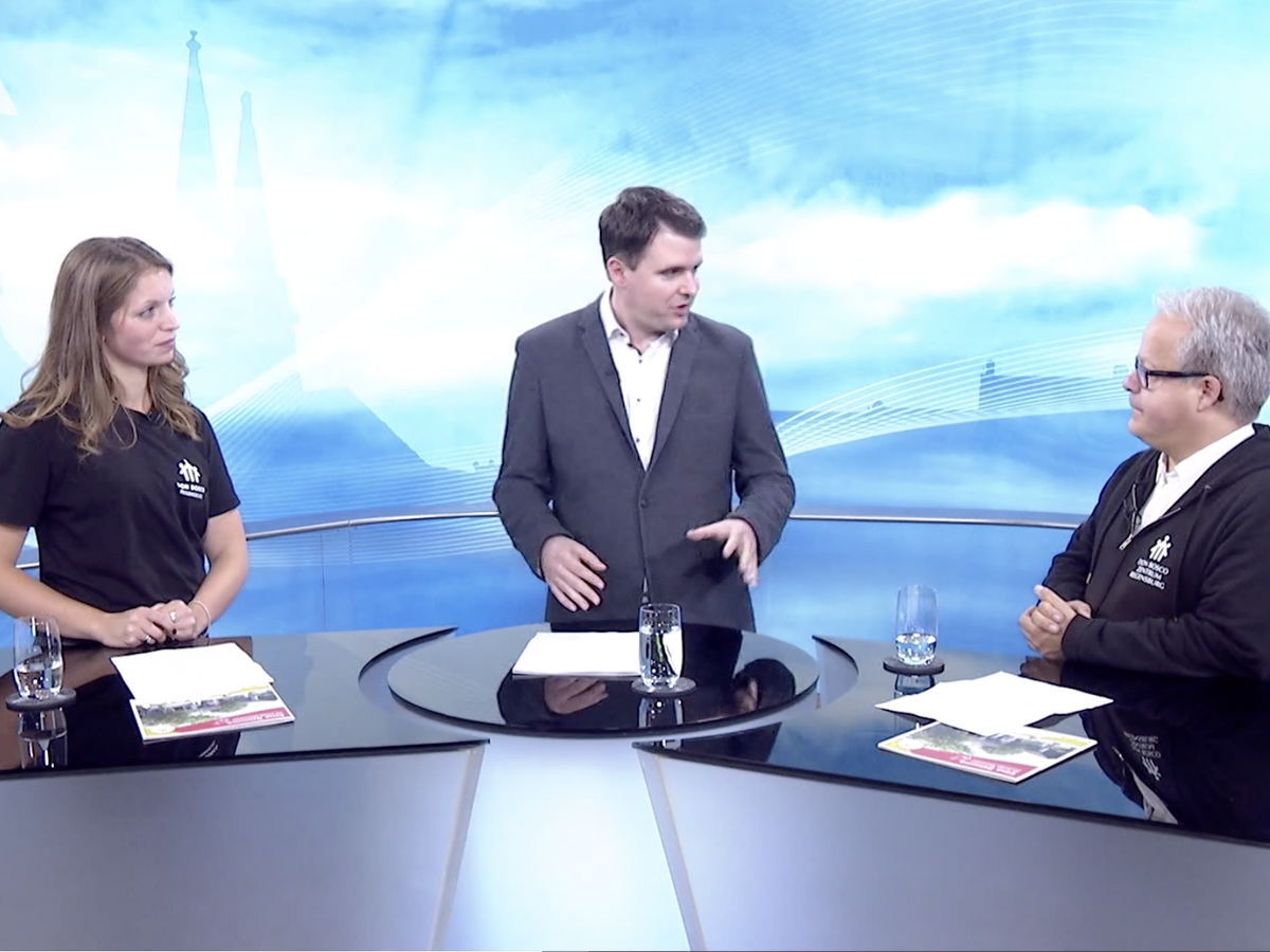 Ein männlicher Moderator interviewt eine Frau und einen anderen Mann in einem TV-Studio, sitzend an einem schwarzen Tisch vor blauem Stadthintergrund.