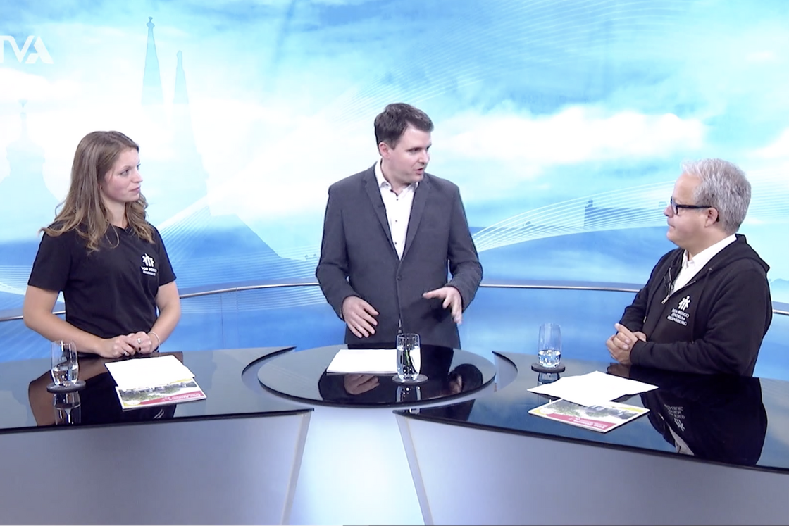 Ein männlicher Moderator interviewt eine Frau und einen anderen Mann in einem TV-Studio, sitzend an einem schwarzen Tisch vor blauem Stadthintergrund.