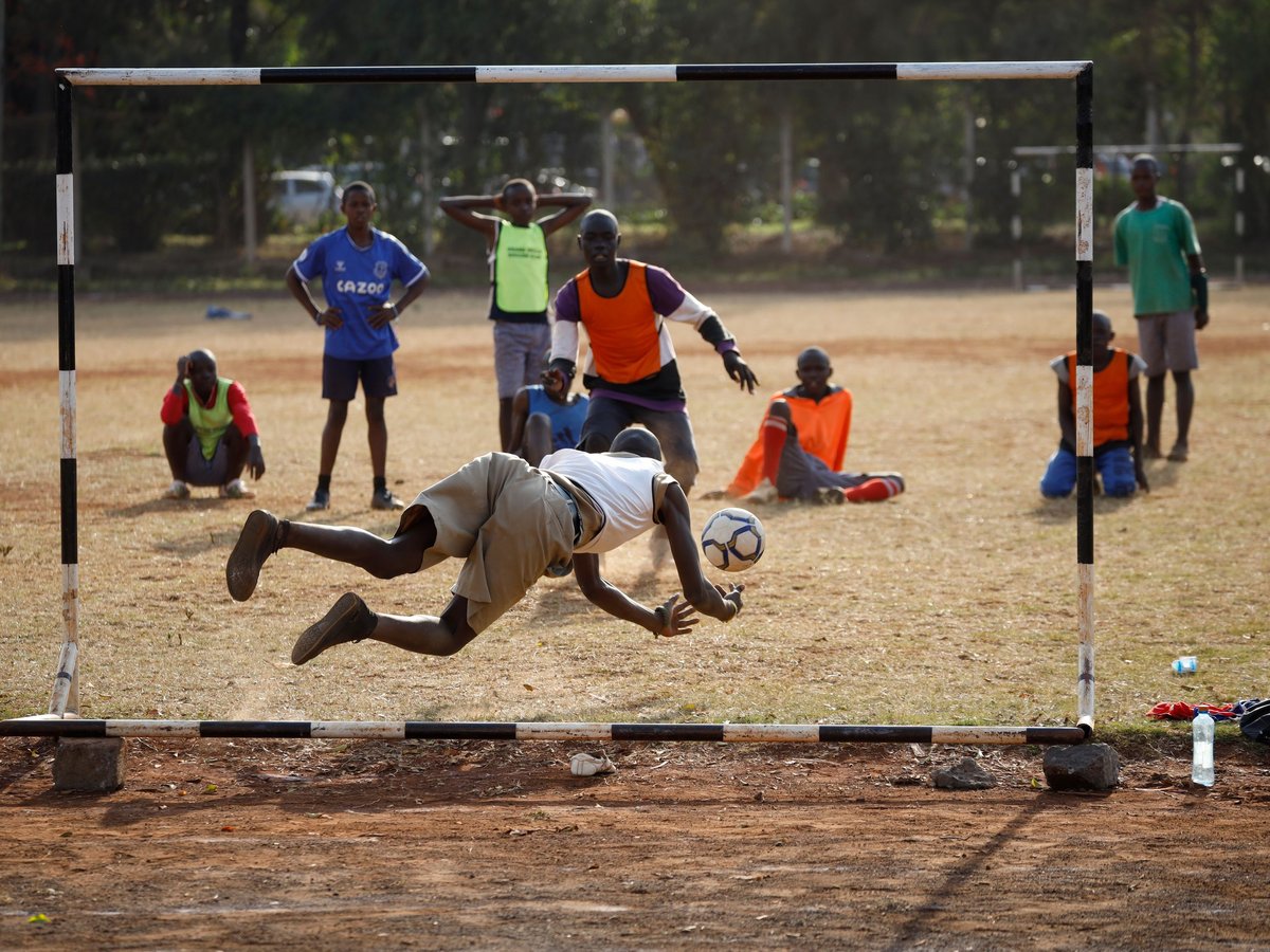 Ein junger Torwart in Shorts und weißem T-Shirt hechtet horizontal nach einem Fußball in einem improvisierten Tor auf staubigem Feld.