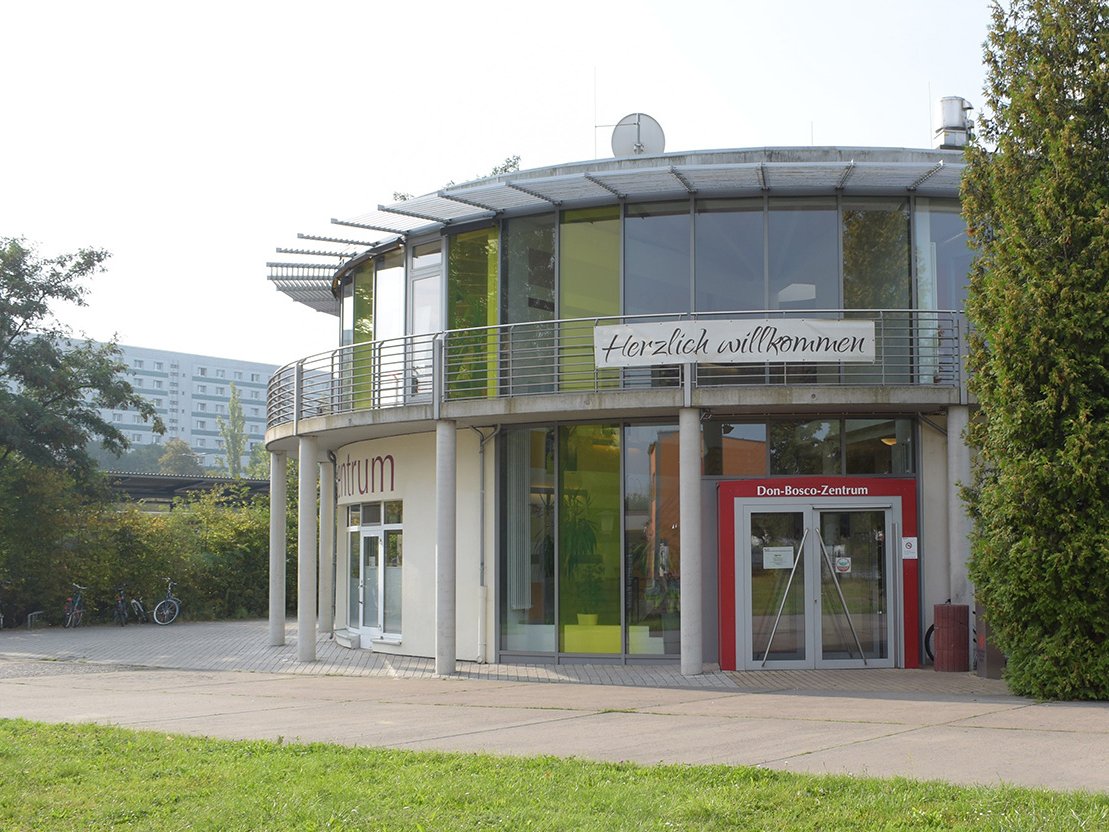 Modernes, rundes Don-Bosco-Zentrum mit großen Fenstern, einem 'Herzlich willkommen'-Banner, Bäumen und einem Wohnhaus.