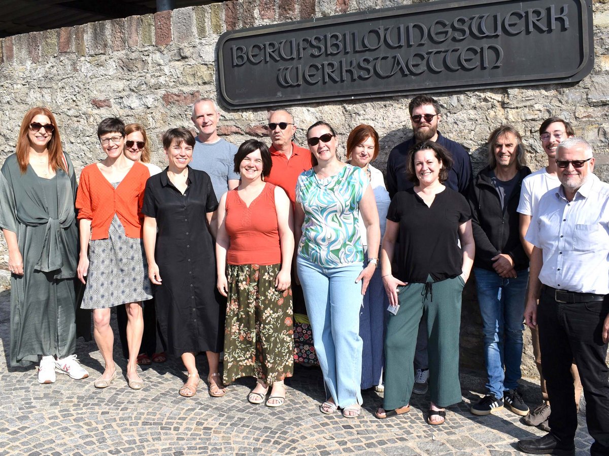 Eine lächelnde Gruppe von etwa 15 Erwachsenen steht vor einer Steinmauer mit dem Schild 'Berufsbildungswerk Werkstätten'.