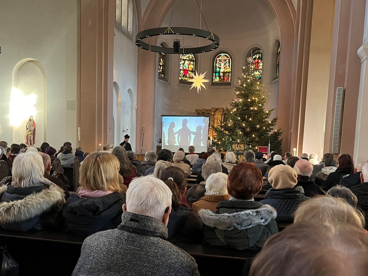 Besucher in einer Kirche schauen auf eine Leinwand mit Silhouetten und einen Geistlichen vor einem geschmückten Weihnachtsbaum.