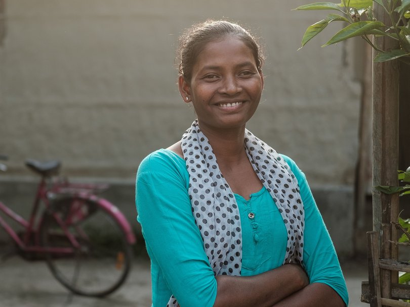 Eine lächelnde junge Frau mit dunkler Haut und einem gepunkteten Schal steht mit verschränkten Armen im Freien, ein verschwommenes rosa Fahrrad im Hintergrund.