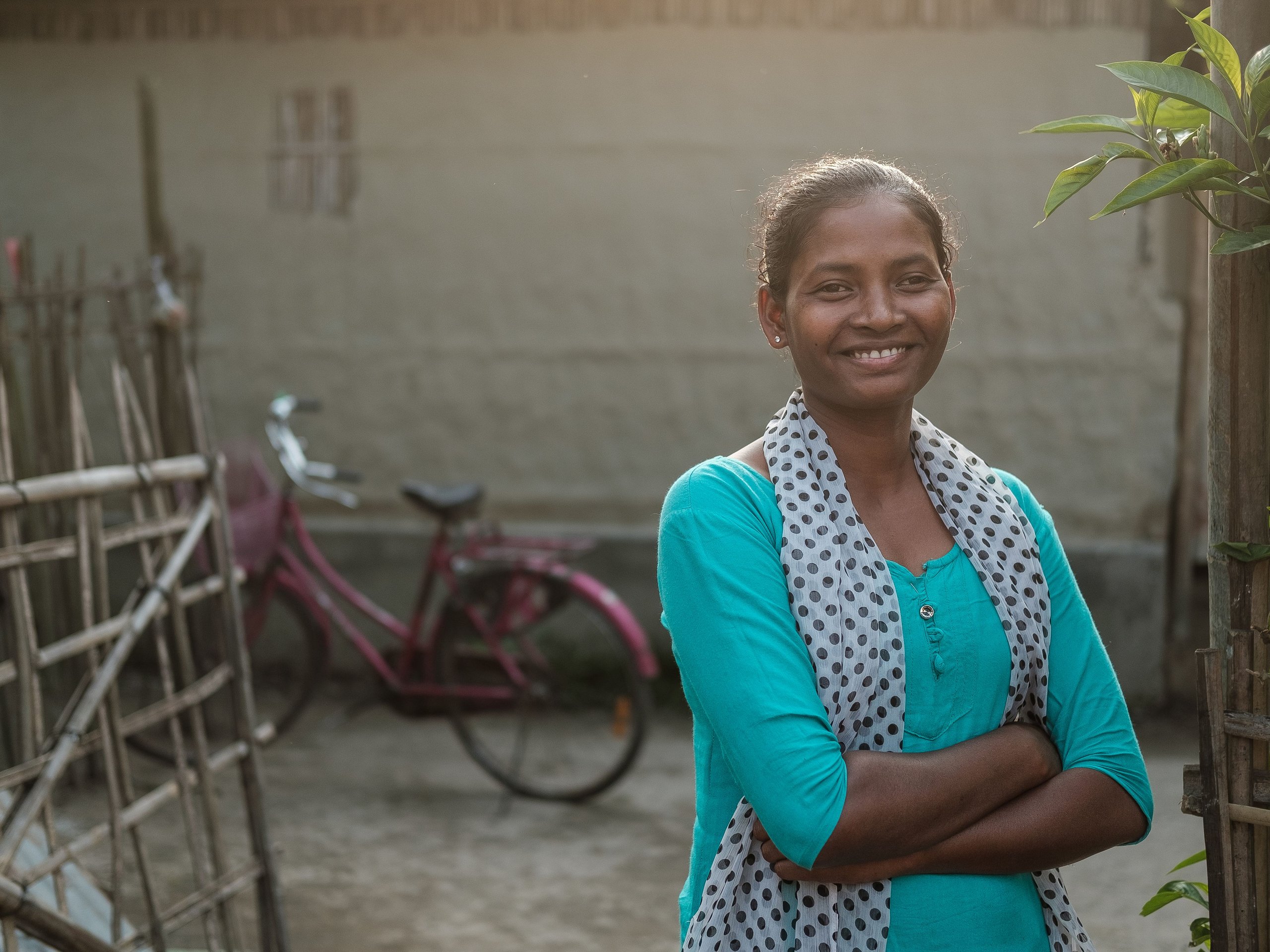 Eine lächelnde junge Frau mit dunkler Haut und einem gepunkteten Schal steht mit verschränkten Armen im Freien, ein verschwommenes rosa Fahrrad im Hintergrund.