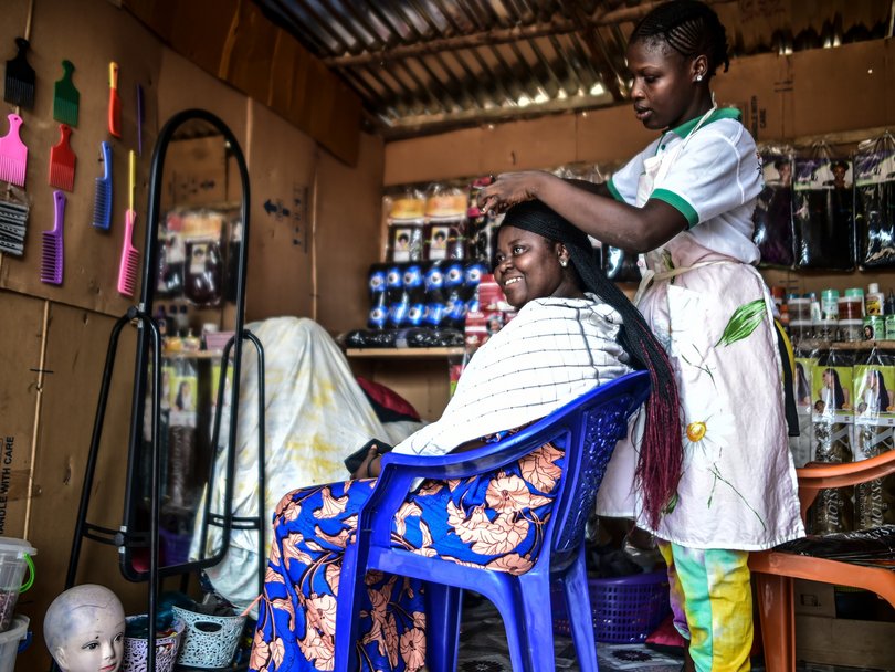 In einem improvisierten Friseursalon flechtet eine Friseurin einer lächelnden Kundin die Haare. Bunte Kämme und Produkte schmücken den Raum.