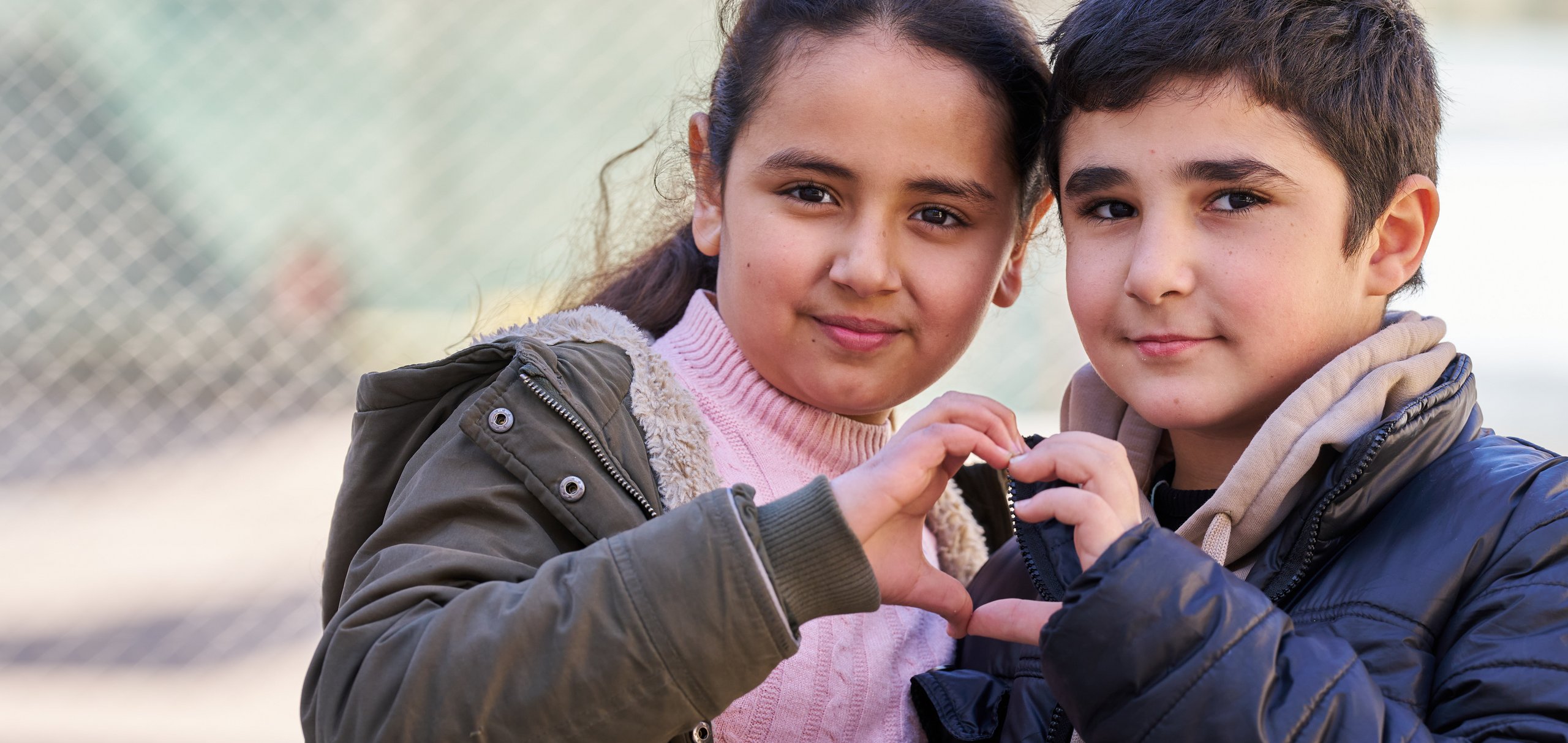 Ein junges Mädchen und ein Junge in Wintermänteln bilden mit ihren Händen ein Herz und schauen in die Kamera.