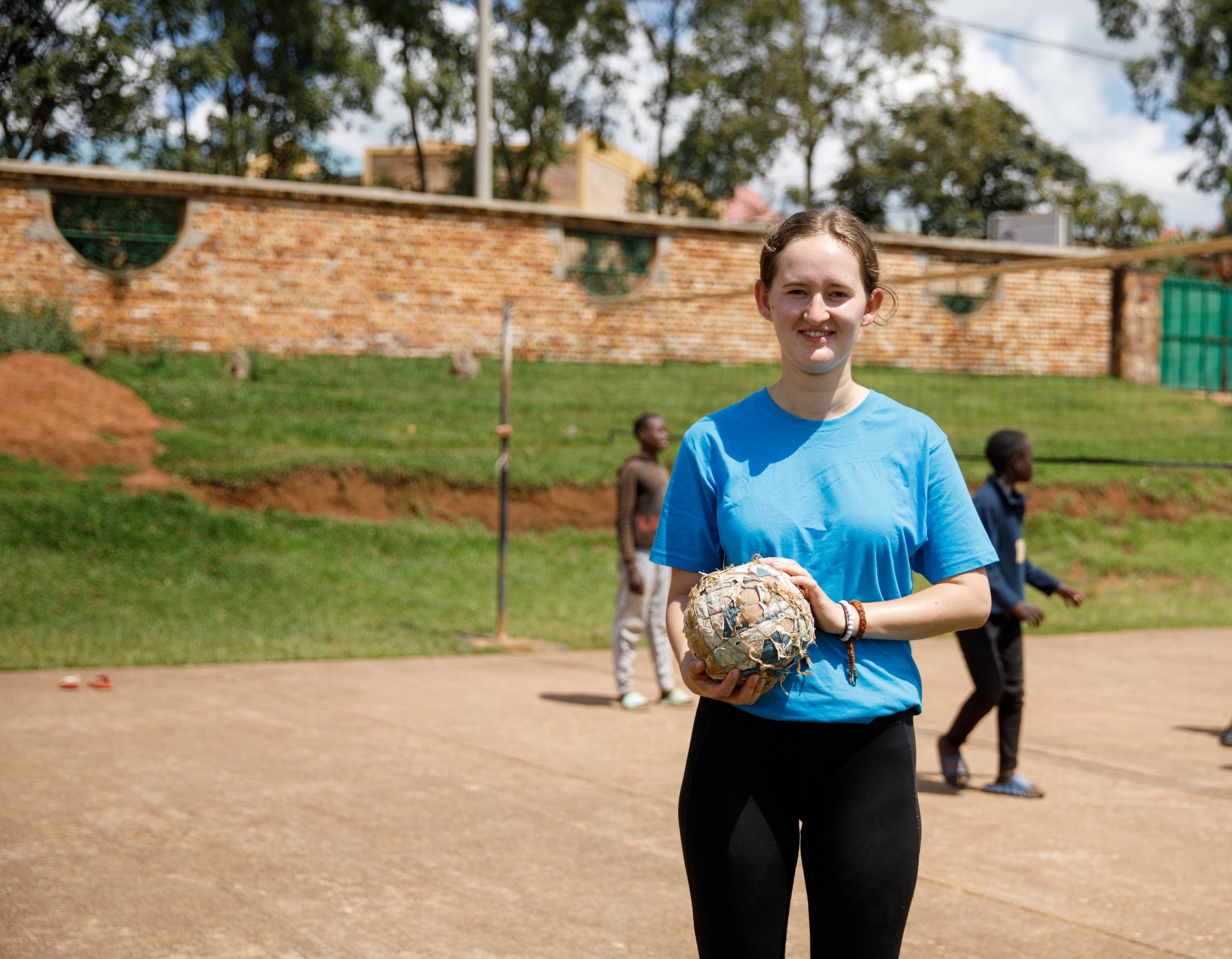 Eine lächelnde junge Frau hält einen selbstgemachten Ball auf einem Sportplatz, während afrikanische Jungen im Hintergrund Volleyball spielen.