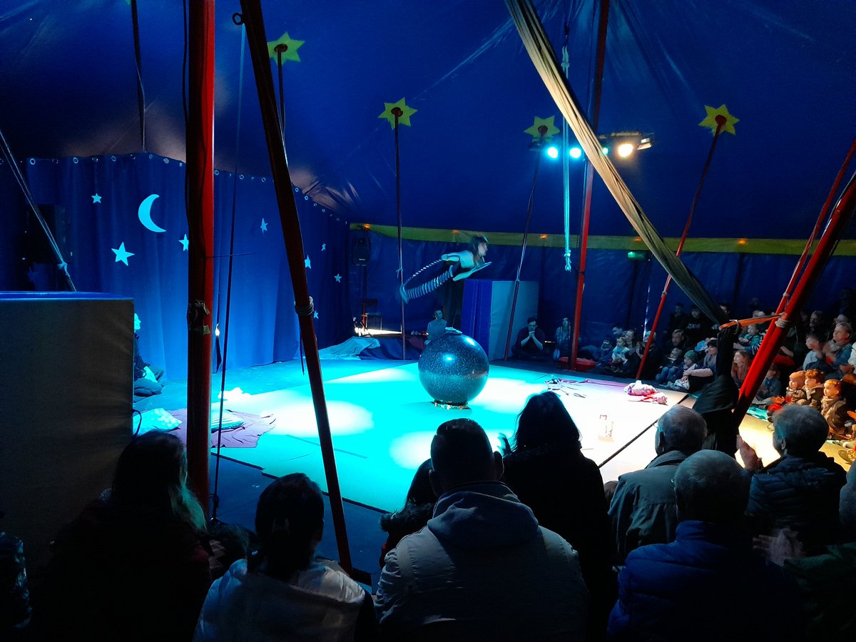 Ein Artist balanciert auf einem großen Ball in einem blau beleuchteten Zirkuszelt vor einem Publikum.