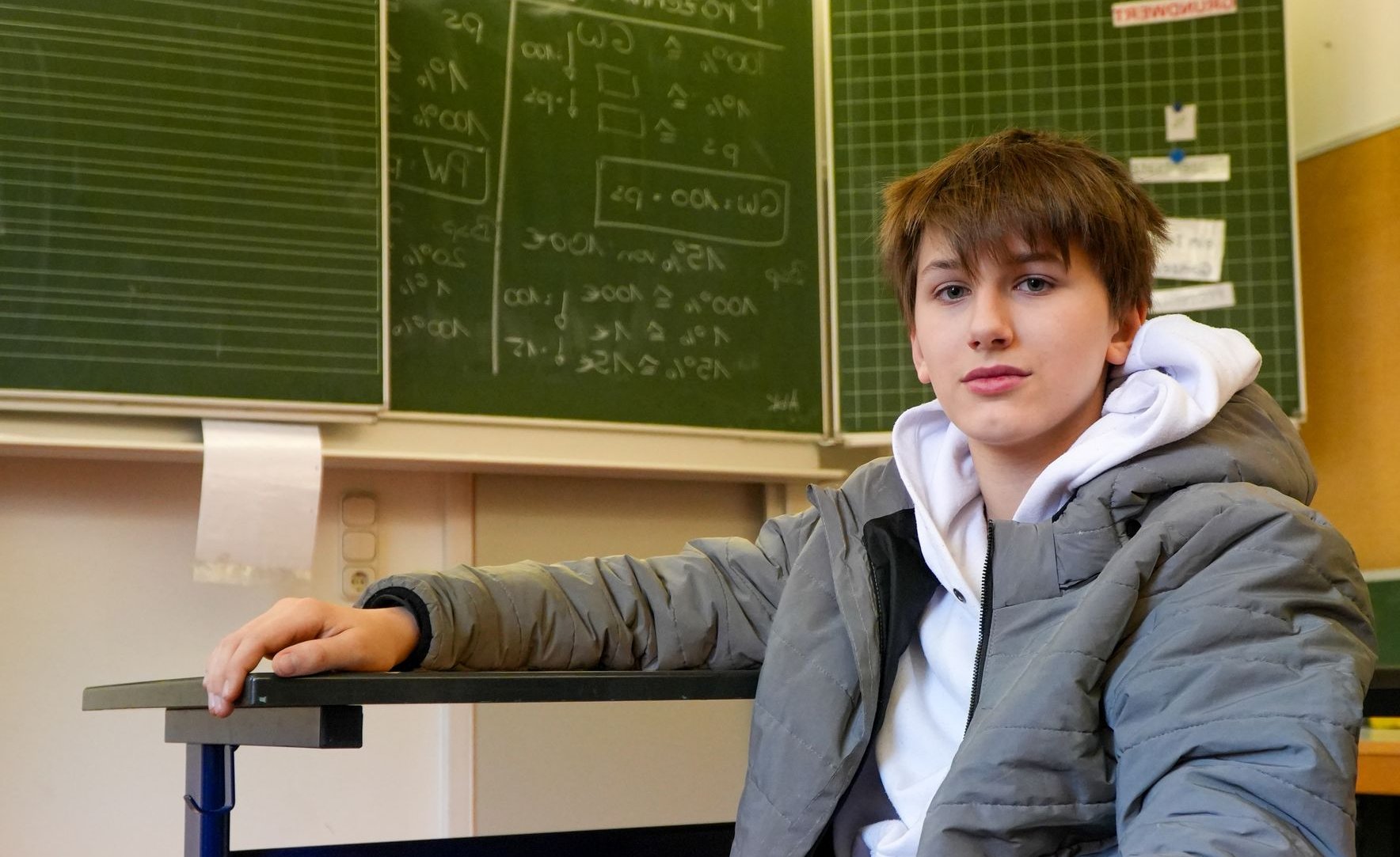 Ein Teenager in grauer Steppjacke und weißem Hoodie sitzt an einem Schultisch und blickt nach vorne. Tafeln im Hintergrund.