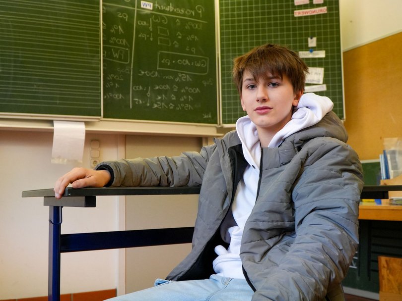 Ein Teenager in grauer Steppjacke und weißem Hoodie sitzt an einem Schultisch und blickt nach vorne. Tafeln im Hintergrund.