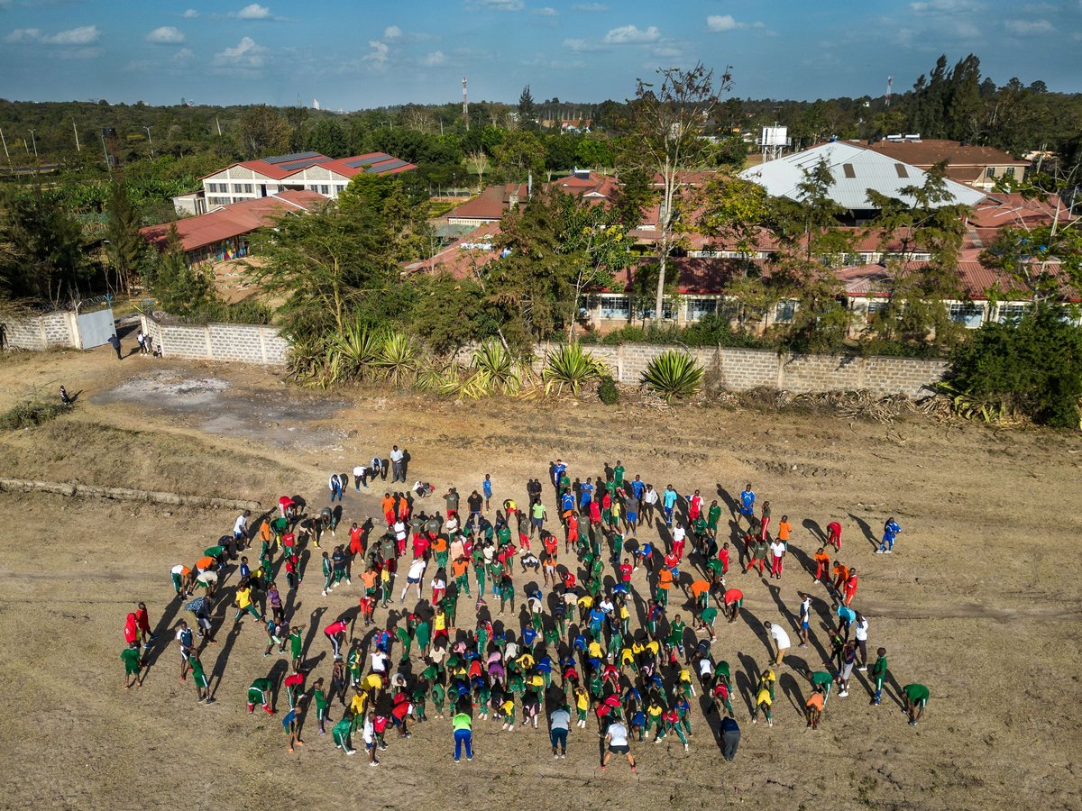 Luftaufnahme: viele junge Menschen in bunter Sportkleidung trainieren auf einem trockenen Feld, mit Gebäuden und Bäumen im Hintergrund.