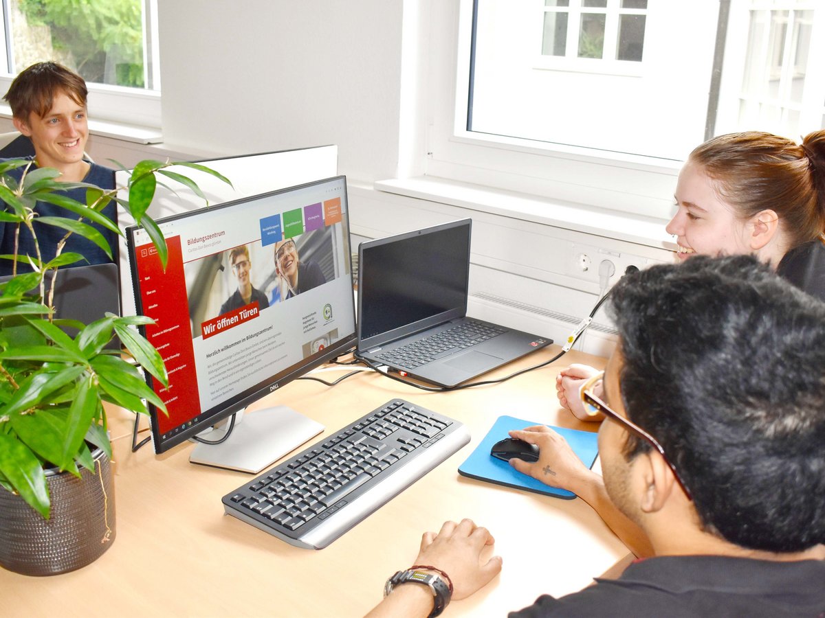 Drei junge Menschen, zwei lächelnd, einer mit der Maus, arbeiten an Computern im Büro; eine Bildungszentrum-Website ist zu sehen.