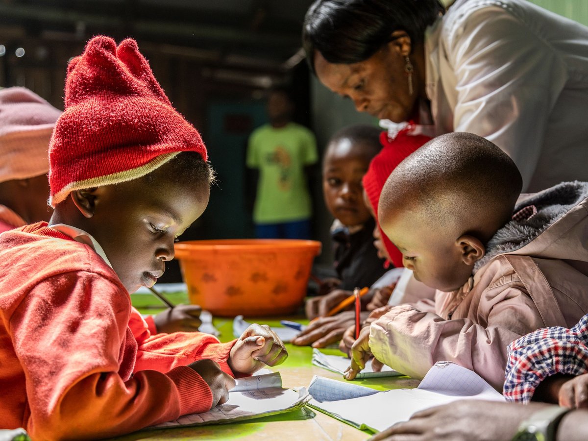 Nahaufnahme von jungen afrikanischen Kindern, darunter eines mit roter Mütze, die aufmerksam an einem Tisch in Notizbücher schreiben, während eine Erwachsene beaufsichtigt.