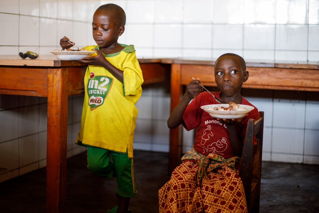 Zwei junge afrikanische Kinder essen eine Mahlzeit aus Reis und Bohnen in einem einfachen Raum mit Fliesenwänden und Holztischen.