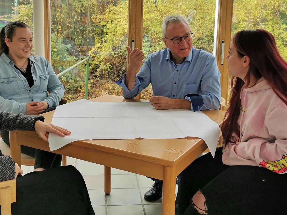 Ein älterer Mann und drei junge Frauen sitzen an einem Tisch mit einem großen weißen Blatt Papier und führen eine lebhafte Diskussion.