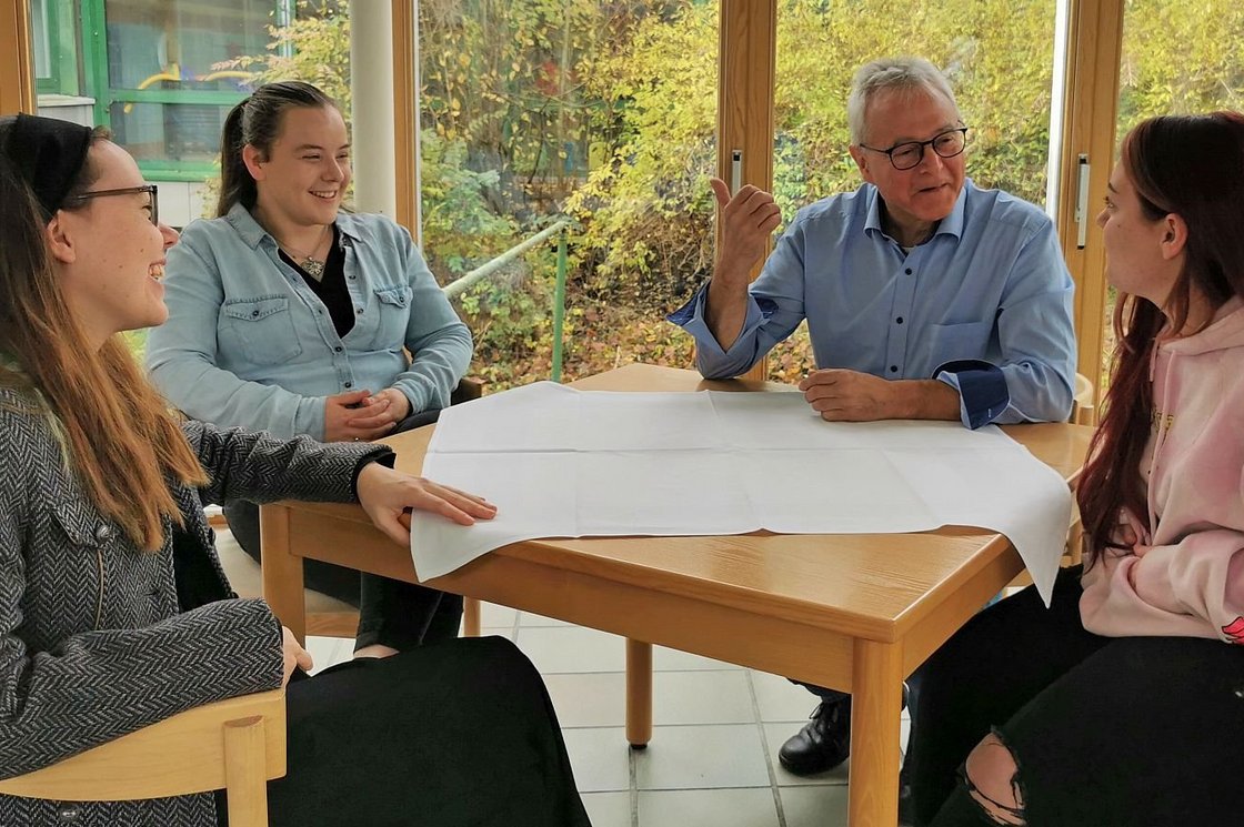 Ein älterer Mann und drei junge Frauen sitzen an einem Tisch mit einem großen weißen Blatt Papier und führen eine lebhafte Diskussion.