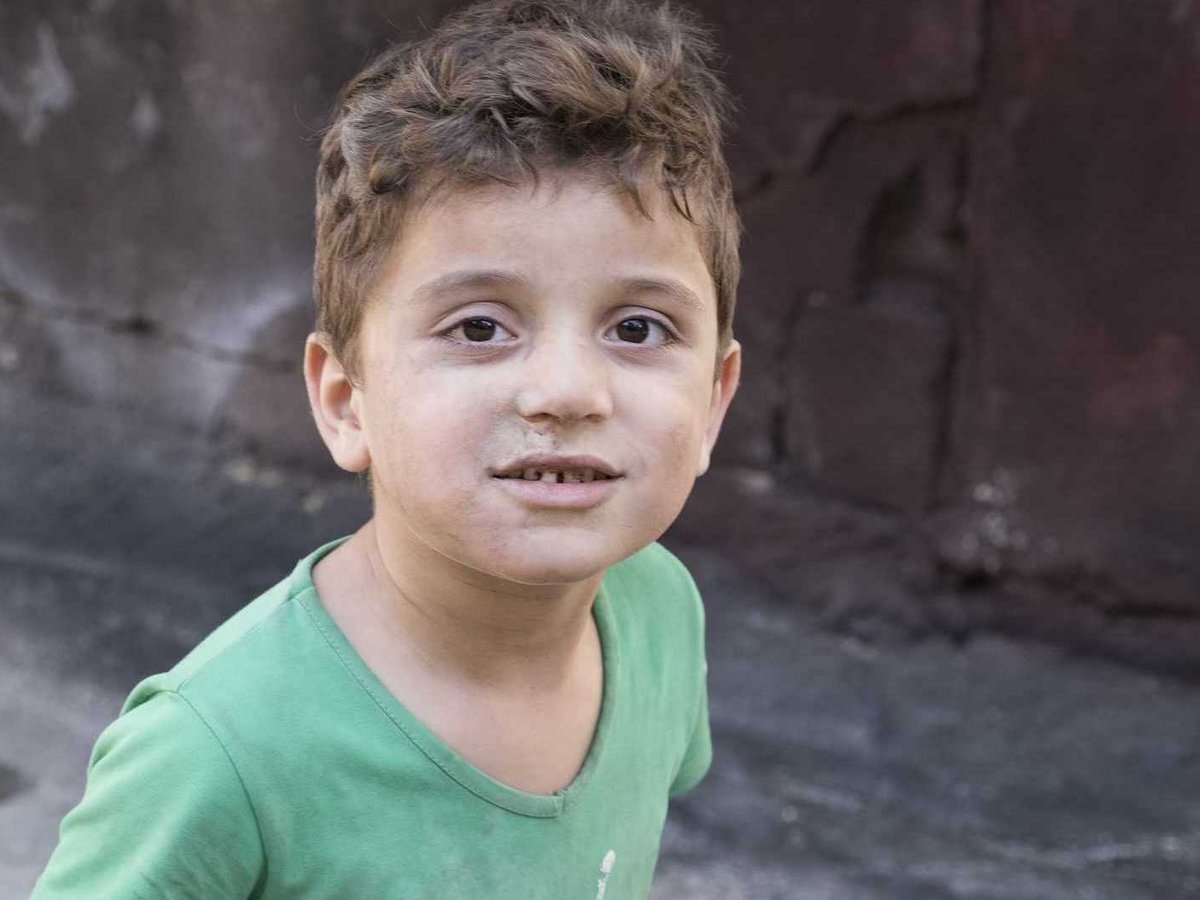 Porträt eines Jungen mit lockigem braunem Haar, schmutzigem Gesicht und fehlenden Vorderzähnen, in grünem Hemd.