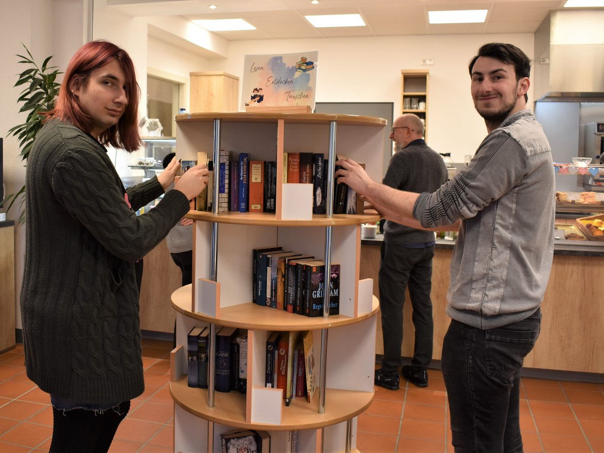 Zwei junge Leute lächeln und ordnen Bücher an einem runden Regal in einer Bücherei oder einem Café.