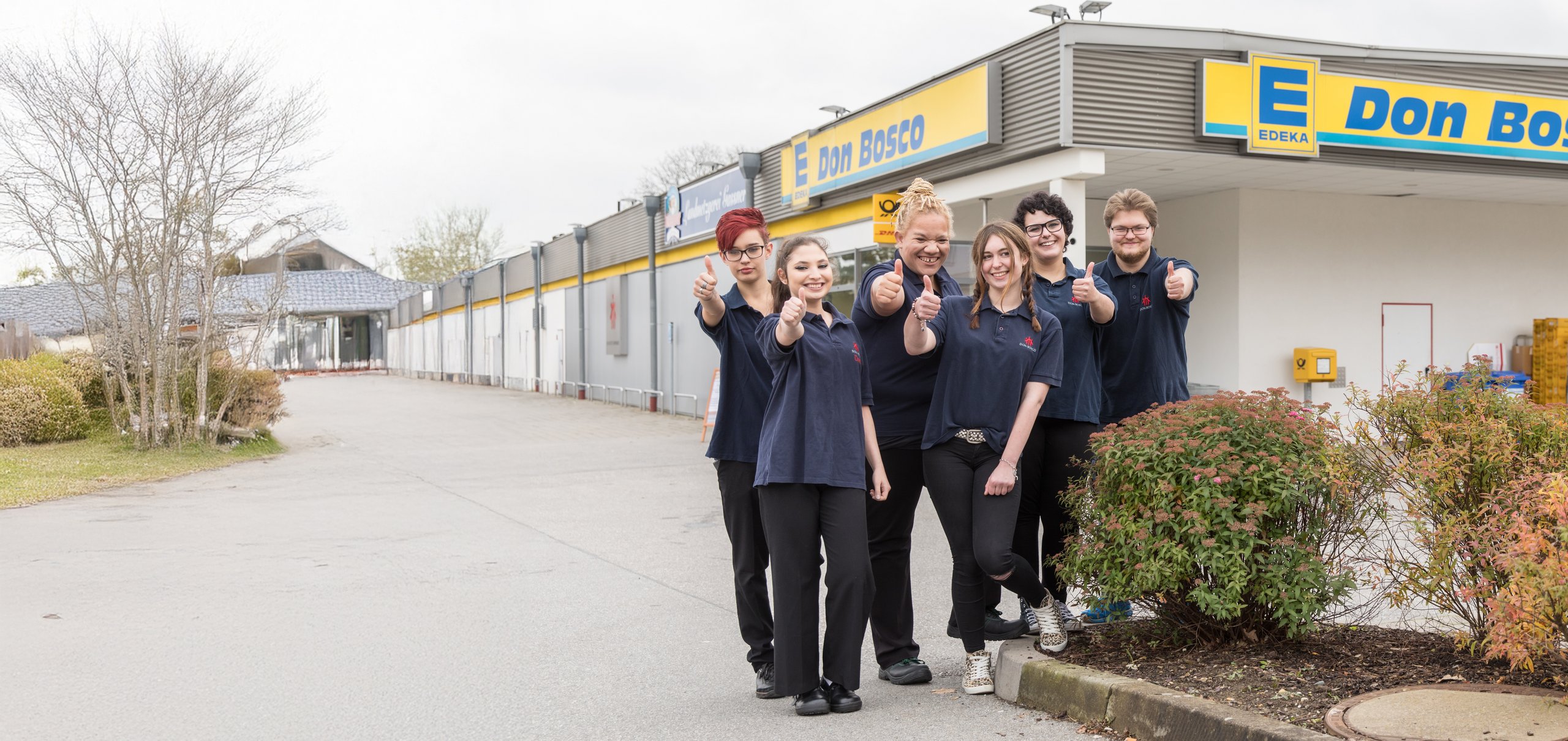 Sechs lächelnde EDEKA Don Bosco Mitarbeiter zeigen vor ihrem Supermarkt Daumen hoch.