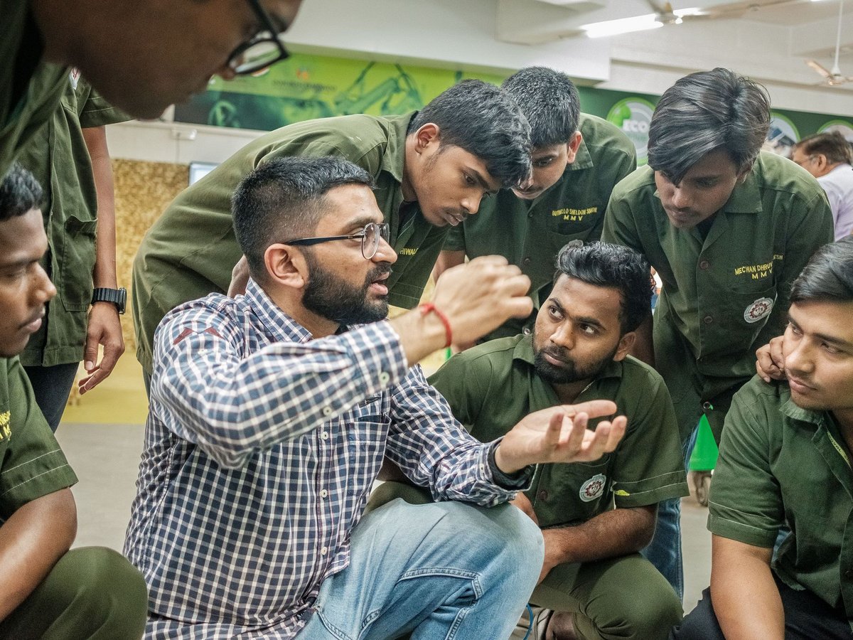 Ein bärtiger Mann im karierten Hemd erklärt einer Gruppe junger Männer in grünen Uniformen etwas, die aufmerksam zuhören.