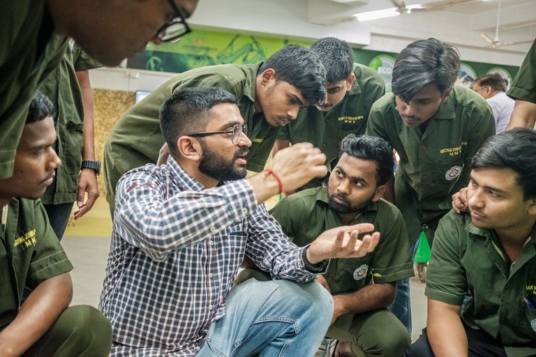 Ein bärtiger Mann im karierten Hemd erklärt einer Gruppe junger Männer in grünen Uniformen etwas, die aufmerksam zuhören.