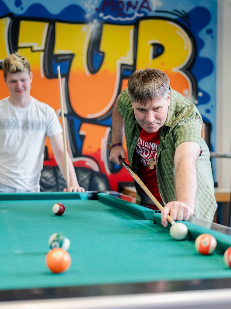 Zwei Männer spielen Billard. Ein Mann beugt sich über den Tisch und stößt mit dem Queue eine Kugel. Im Hintergrund bunte Graffiti.