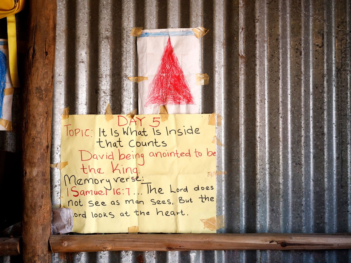 Ein gelbes Poster mit einem Bibelvers über David und Kinderzeichnungen an einer Wellblechwand.