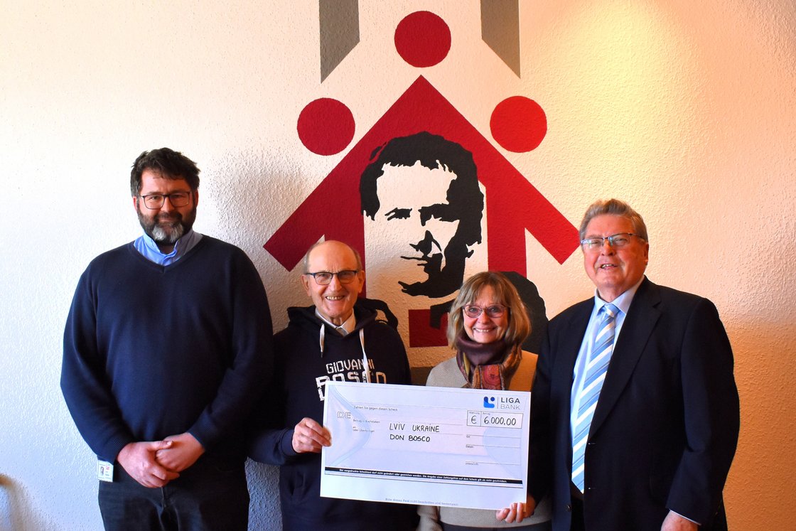 Vier Personen, zwei halten einen Scheck über 6.000 € für Lemberg, Ukraine, Don Bosco, vor Wandgrafik.