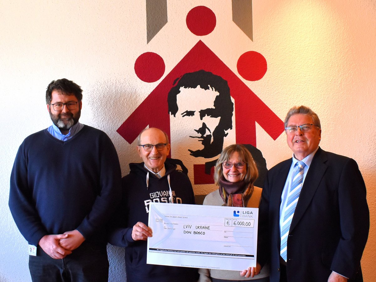 Vier Personen, zwei halten einen Scheck über 6.000 € für Lemberg, Ukraine, Don Bosco, vor Wandgrafik.