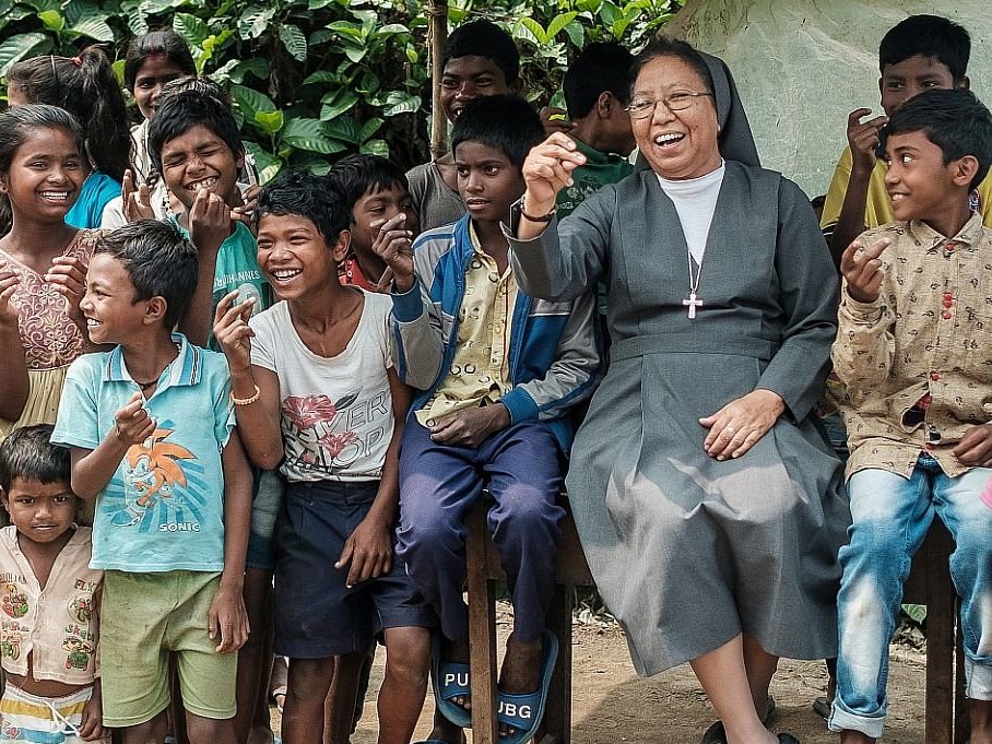 Eine lachende Nonne sitzt inmitten einer großen Gruppe fröhlicher Kinder in einer ländlichen Umgebung.