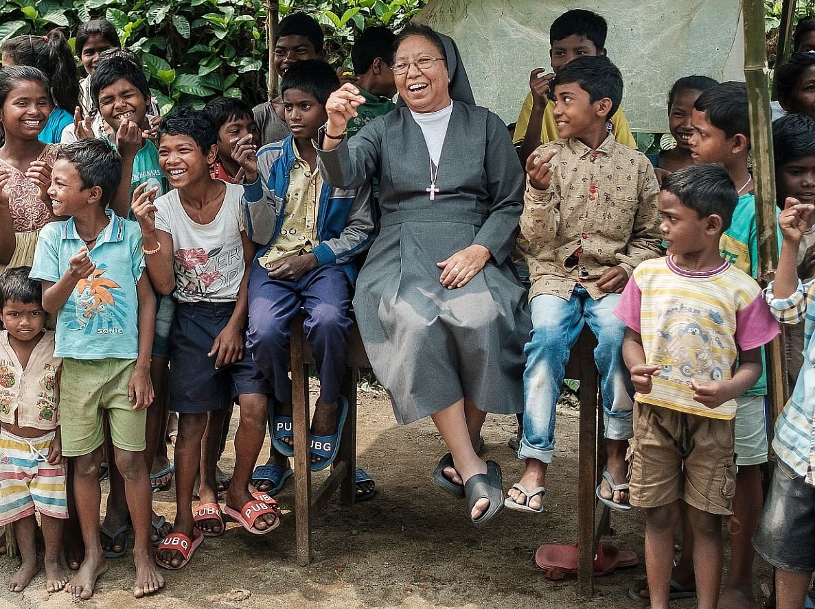 Eine lachende Nonne sitzt inmitten einer großen Gruppe fröhlicher Kinder in einer ländlichen Umgebung.