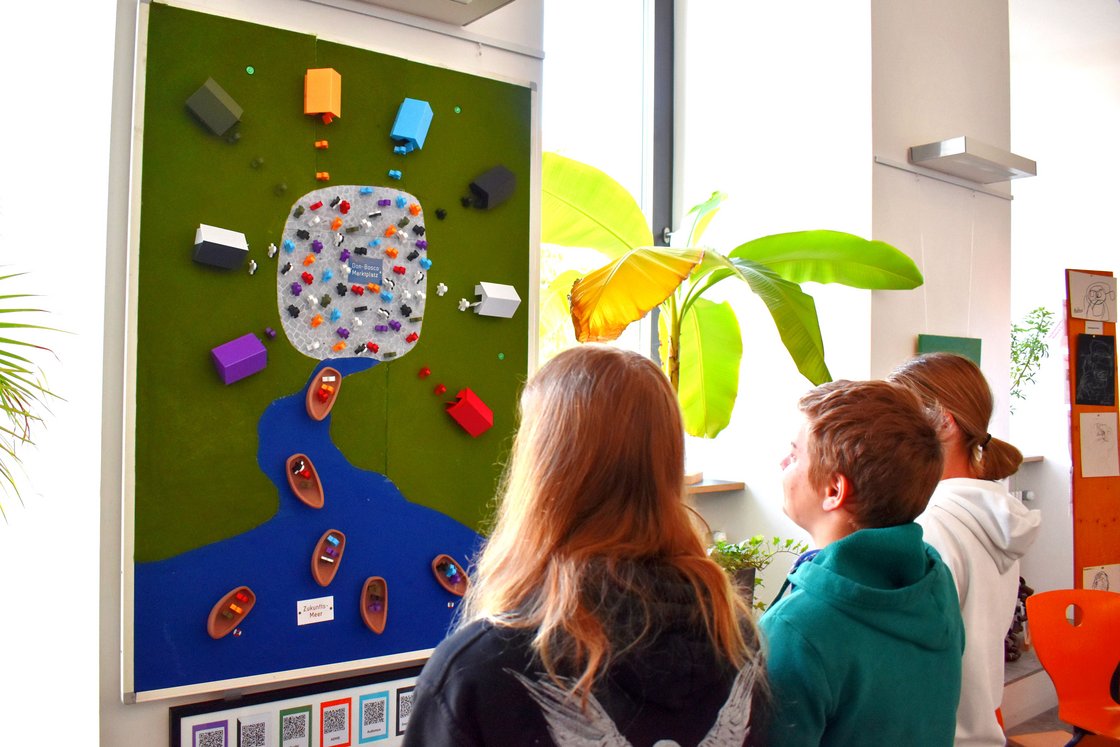 Drei Personen betrachten eine interaktive Tafel mit einem Modell von Häusern, Figuren und Booten auf Fluss/Meer.