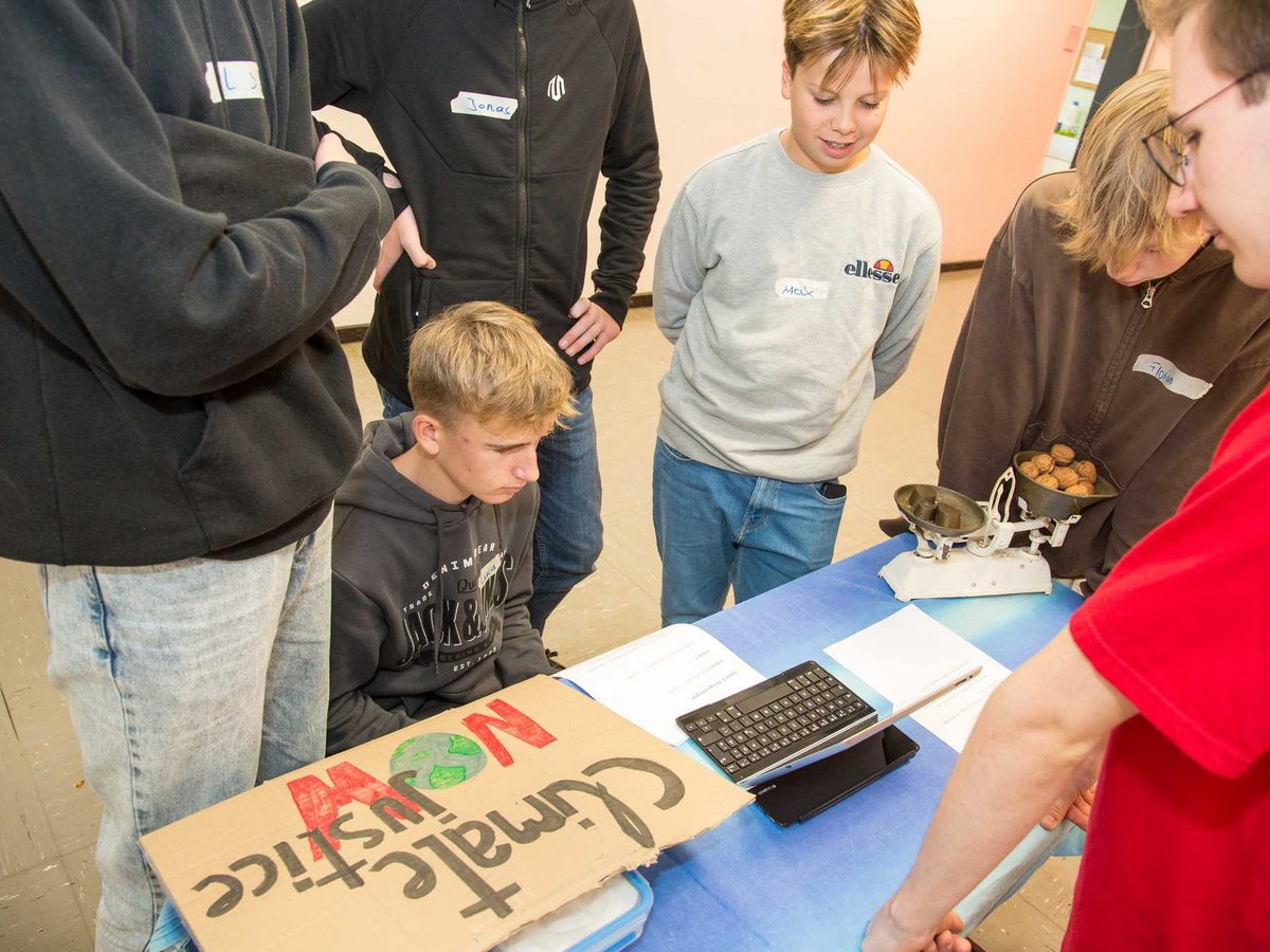 Schüler um einen Tisch mit 'Klimagerechtigkeit'-Schild, Waage mit Walnüssen und Taschenrechner bei einem Projekt.