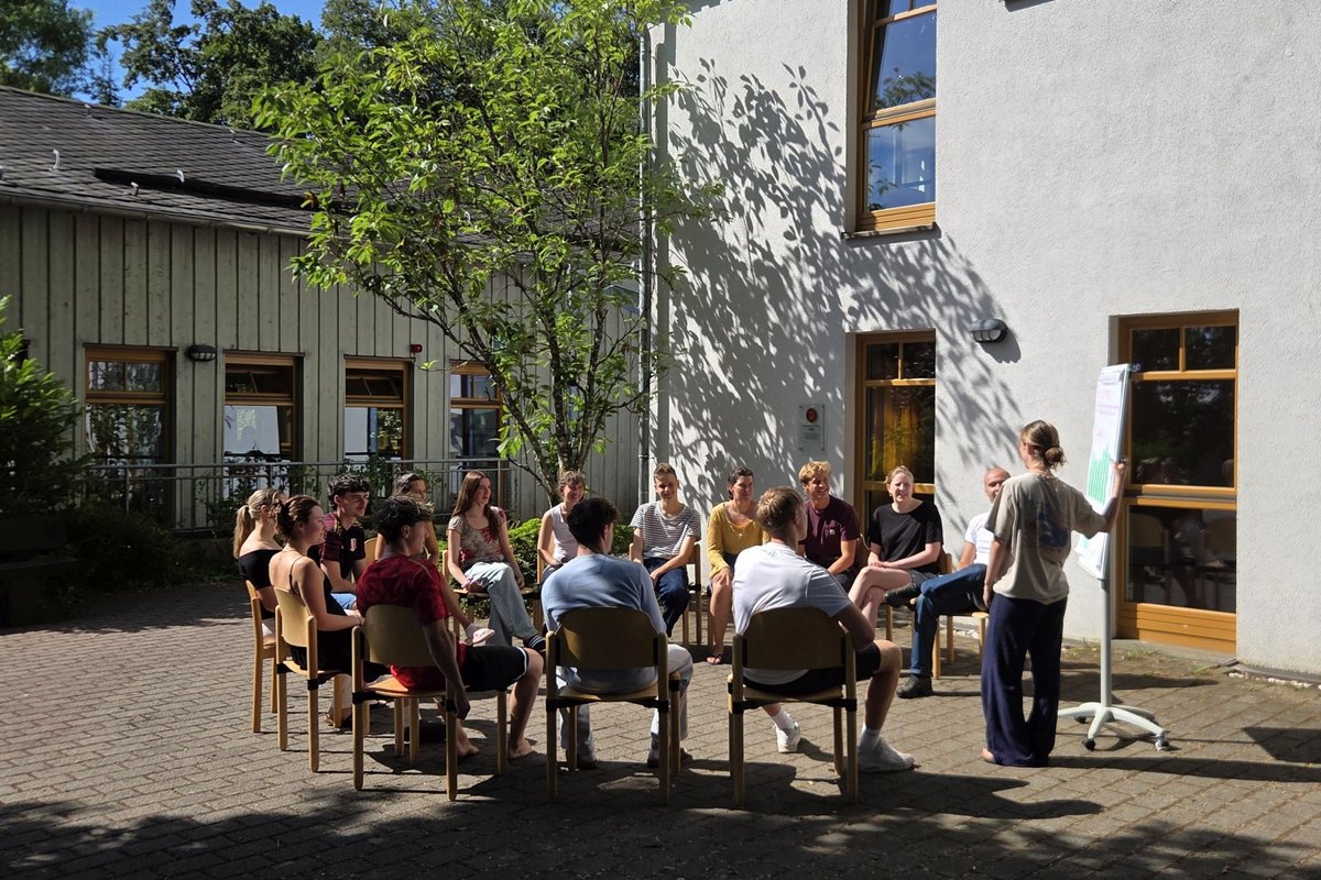 Gruppe junger Menschen im Stuhlkreis vor Gästehaus mit Flipchart und Baum
