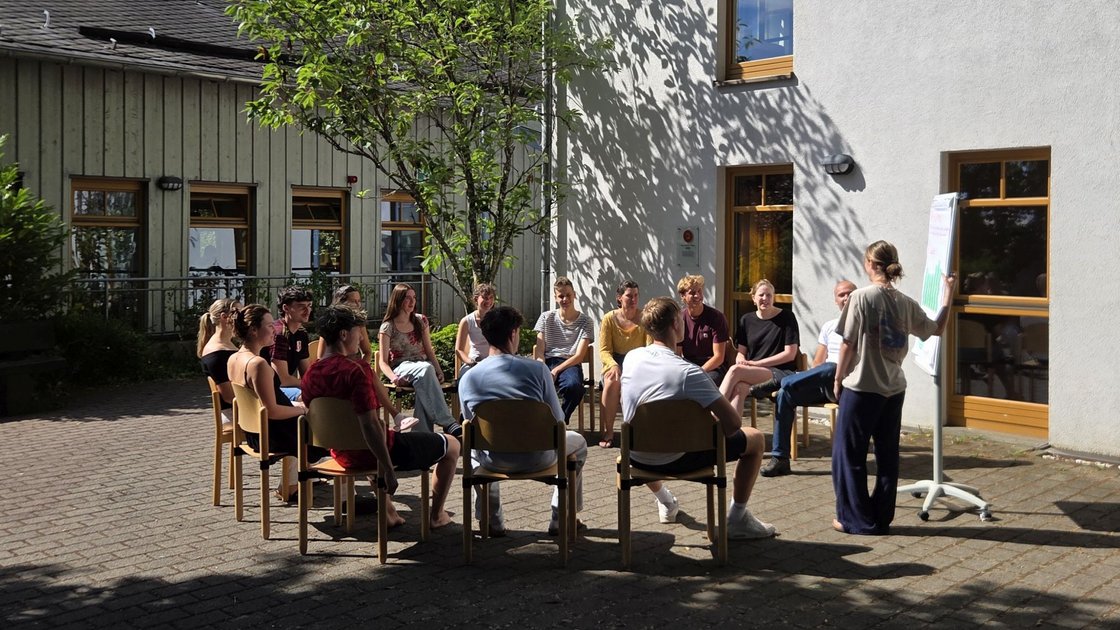 Gruppe junger Menschen im Stuhlkreis vor Gästehaus mit Flipchart und Baum 