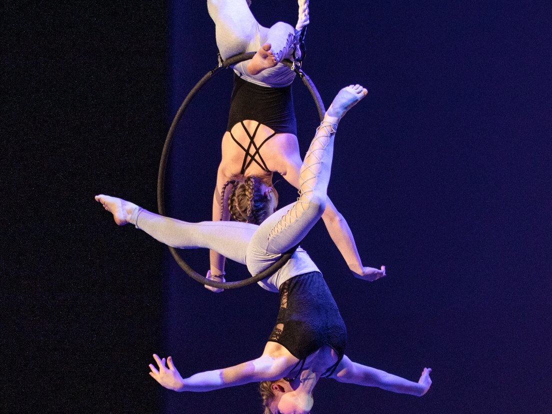 Zwei Luftakrobatinnen führen eine umgekehrte Pose an einem Lyra-Reifen vor dunklem Hintergrund aus, eine über der anderen.