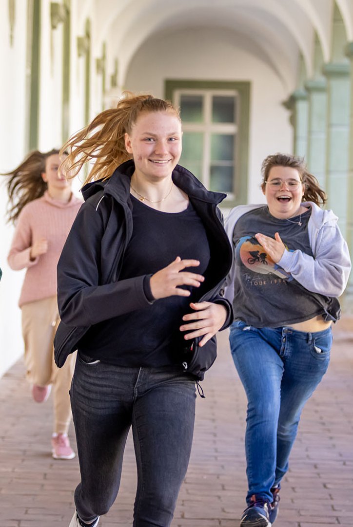 Eine Gruppe lachender junger Leute rennt fröhlich einen langen, gewölbten Gang mit Säulen entlang.