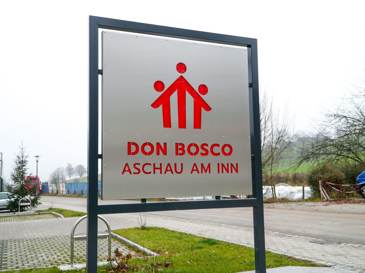 Außenschild für Don Bosco Aschau am Inn mit rotem Logo von drei stilisierten Figuren auf Metallhintergrund.