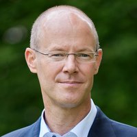 Porträt eines lächelnden Mannes mit kurzen Haaren und Brille in dunklem Anzug und weißem Hemd vor grünem Hintergrund.