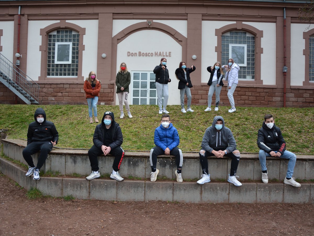 Eine Gruppe von Jugendlichen mit Masken steht und sitzt vor der Don Bosco Halle; einige Mädchen zeigen Daumen nach unten.