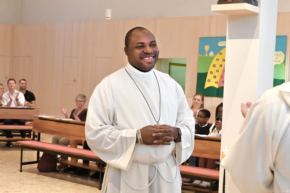 Lächelnder Schwarzer Priester in weißem Gewand in einer Kirche mit klatschenden Gemeindemitgliedern im Hintergrund.