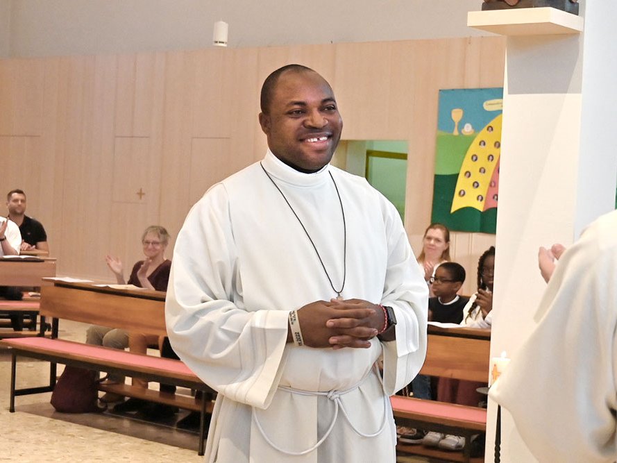 Lächelnder Schwarzer Priester in weißem Gewand in einer Kirche mit klatschenden Gemeindemitgliedern im Hintergrund.