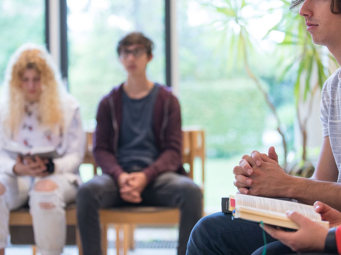 Gruppe junger Erwachsener beim Lernen oder Diskutieren. Vordergrund: Hände mit Buch; Hintergrund: verschwommene Schüler.