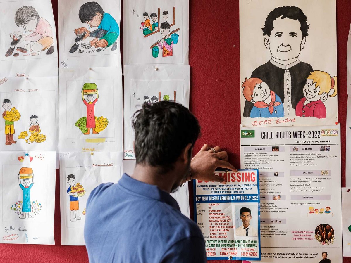 Person pinnt ein Vermisstenplakat an eine rote Pinnwand mit Kinderzeichnungen und einem 'Kinderrechtswoche'-Programm.