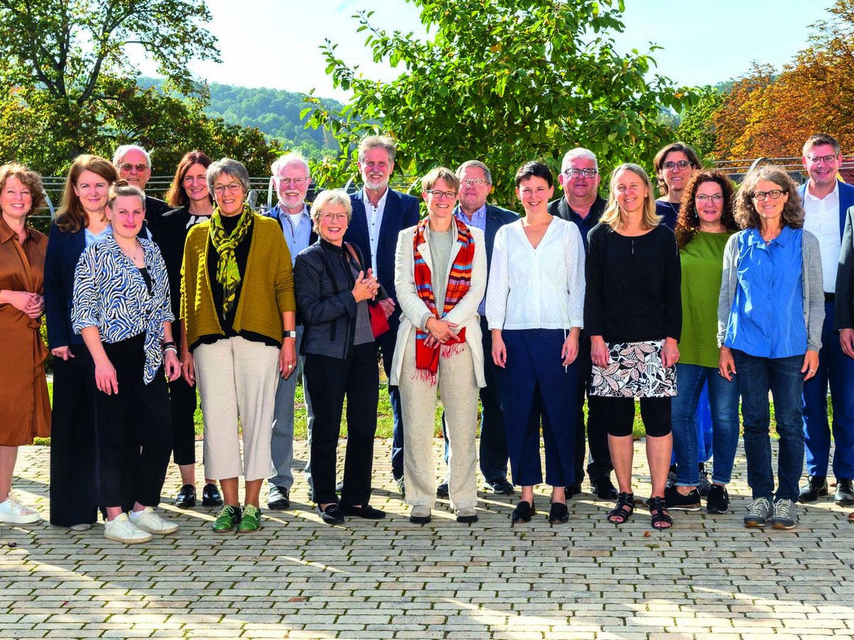 Gruppe von etwa 20 Personen, Männer und Frauen, lächeln in die Kamera, stehen auf gepflastertem Boden im Freien mit Bäumen und Hügeln.