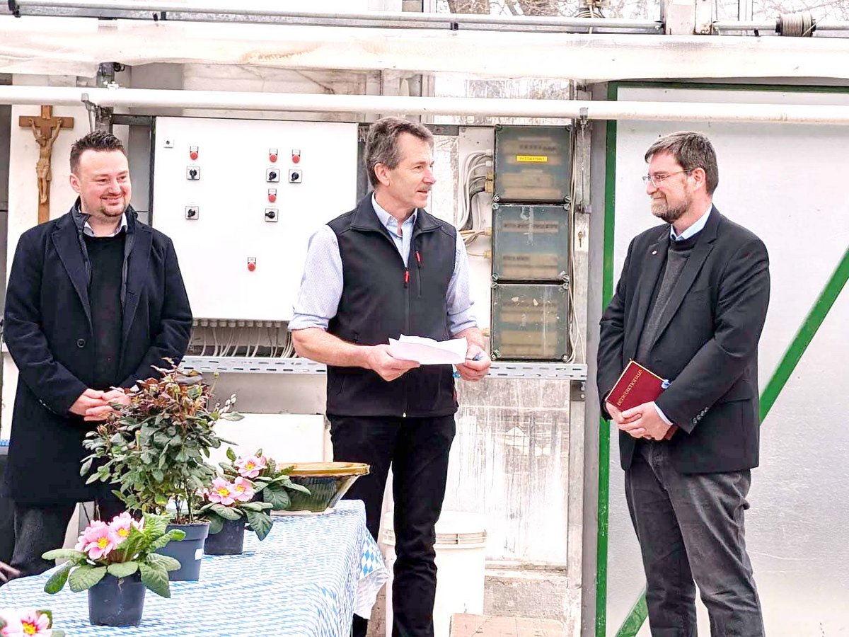 Drei Männer in einem Gewächshaus; einer spricht, Papiere haltend. Ein Tisch mit rosa Topfblumen steht im Vordergrund.