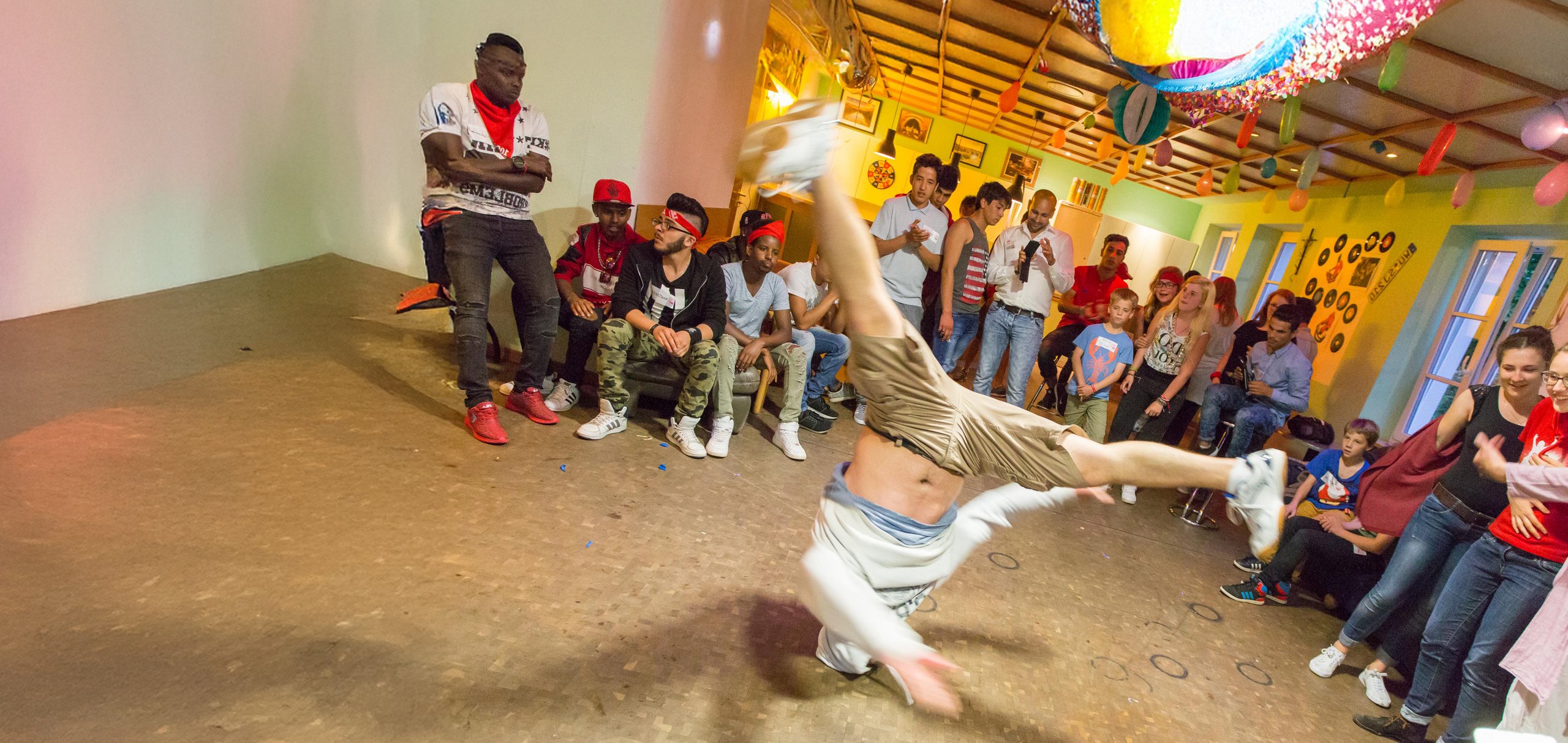 Ein Breakdancer führt einen akrobatischen Handstand-Move vor einer jubelnden Menge in einem festlich geschmückten Raum auf.